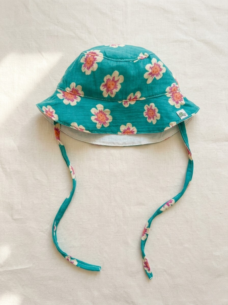 A product flat-lay photograph of the Joy collection baby bucket hat displayed on a neutral cream linen background. The hat is crafted from vibrant teal or turquoise fabric featuring an all-over pink, orange, and cream floral print, complete with matching chin tie straps and a small woven brand logo on the brim edge.