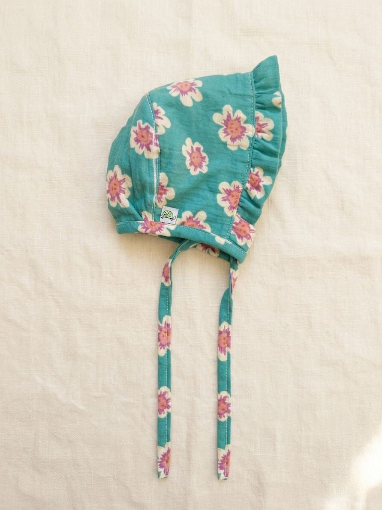 A product flat-lay photograph of a baby muslin bonnet shown from a side profile against a soft off-white linen background. Made from vibrant teal cotton fabric with a pink and cream floral print, the bonnet features a delicate ruffled brim, long matching chin ties extending downward, and a small woven logo tag near the side seam.