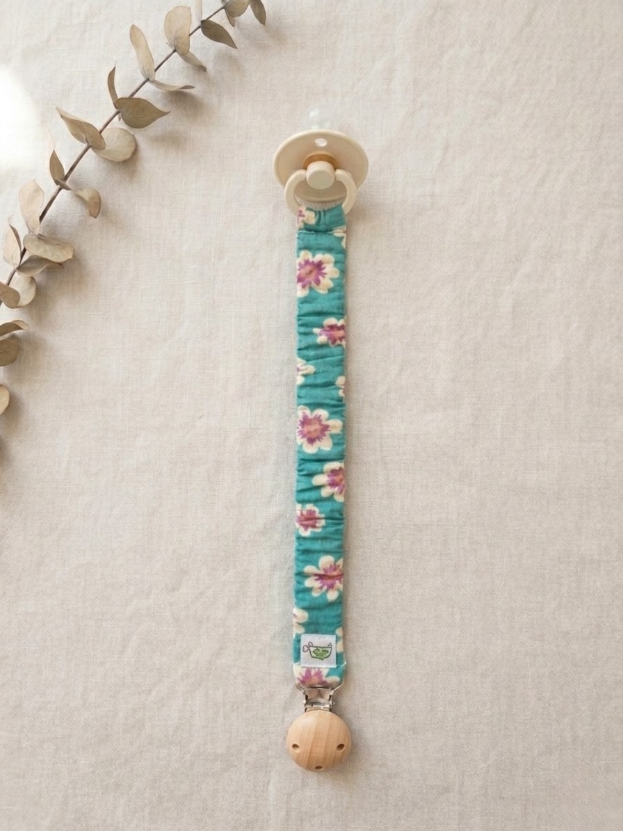 A product flat-lay photograph of a baby pacifier clip with a bright teal fabric sleeve adorned in a pink and yellow floral print, displayed vertically on a textured off-white linen background. The clip features a round natural wooden clasp at the bottom with a small woven brand logo tag, and a cream-colored baby pacifier attached to the top loop. A dried eucalyptus sprig accents the upper left corner of the frame.