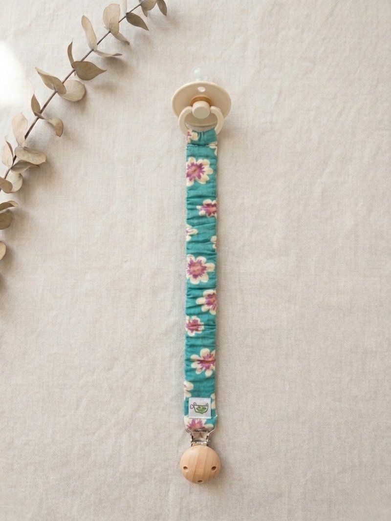 A product flat-lay photograph of a baby pacifier clip with a bright teal fabric sleeve adorned in a pink and yellow floral print, displayed vertically on a textured off-white linen background. The clip features a round natural wooden clasp at the bottom with a small woven brand logo tag, and a cream-colored baby pacifier attached to the top loop. A dried eucalyptus sprig accents the upper left corner of the frame.
