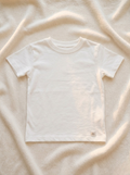 Handmade white baby T-shirt in soft breathable cotton, minimalist short-sleeve design by La Tortuga Marisa, laid on neutral textured fabric background
