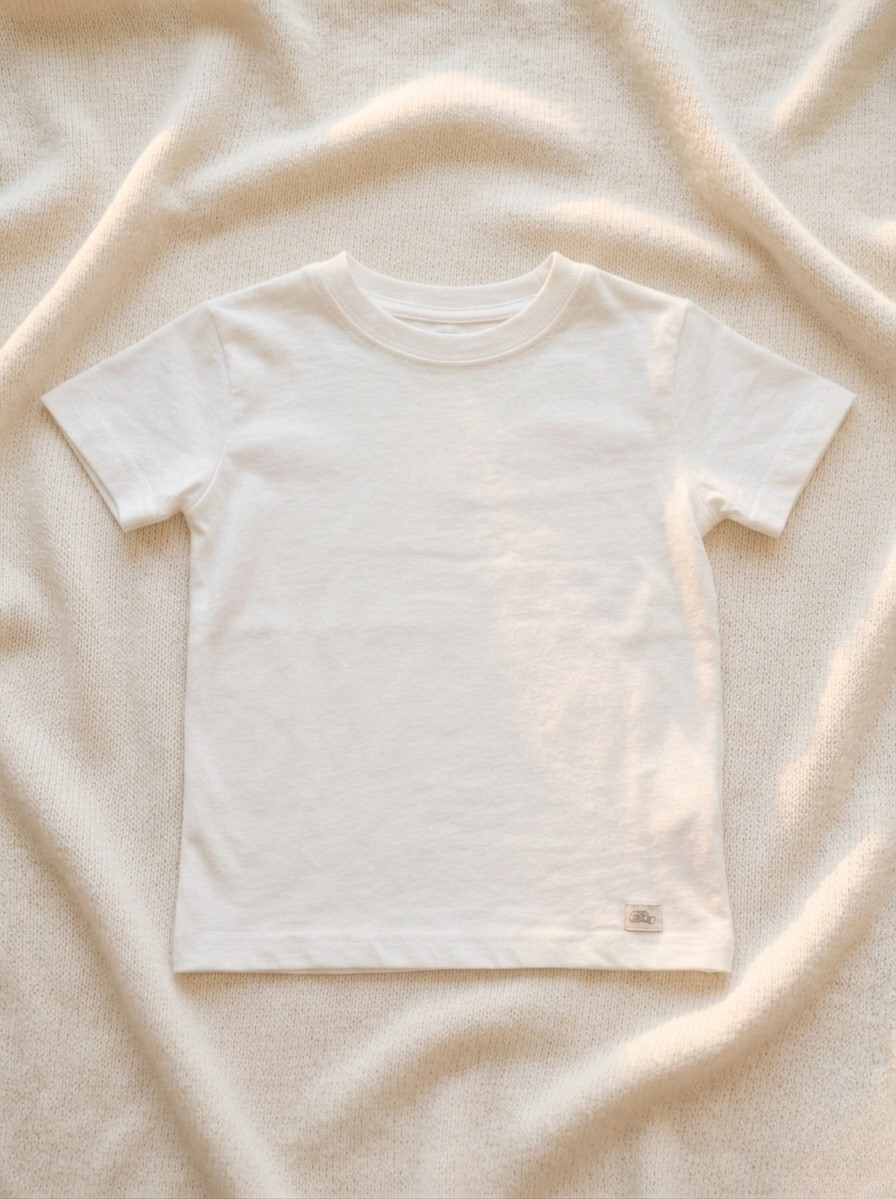 Handmade white baby T-shirt in soft breathable cotton, minimalist short-sleeve design by La Tortuga Marisa, laid on neutral textured fabric background