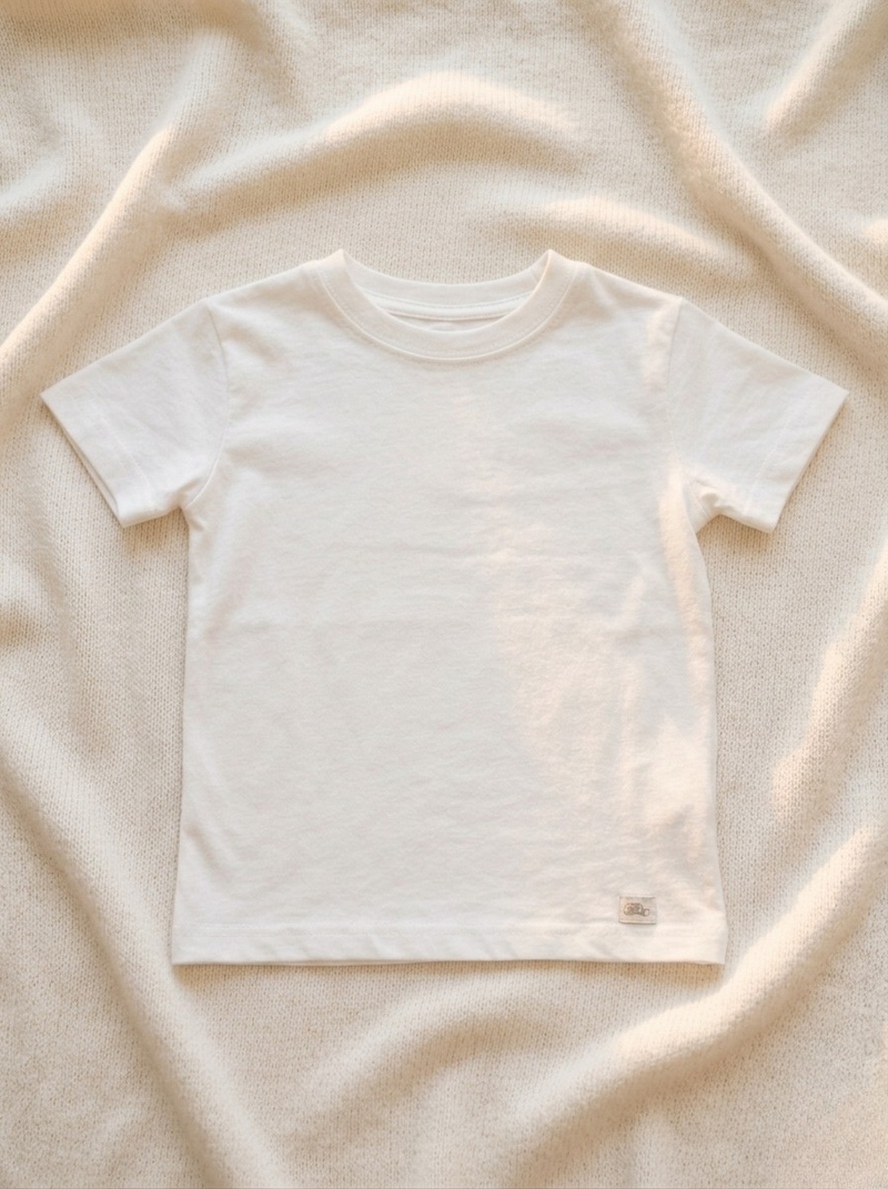 Handmade white baby T-shirt in soft breathable cotton, minimalist short-sleeve design by La Tortuga Marisa, laid on neutral textured fabric background