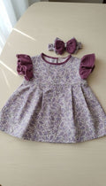 A flat lay photograph of the Larisa baby dress and matching headband. The dress features a lavender and purple paisley print body with plum-colored ruffled muslin sleeves and a matching plum neckline trim. The coordinating headband has a large plum muslin bow with a paisley center, all displayed on a light-colored wooden surface.
