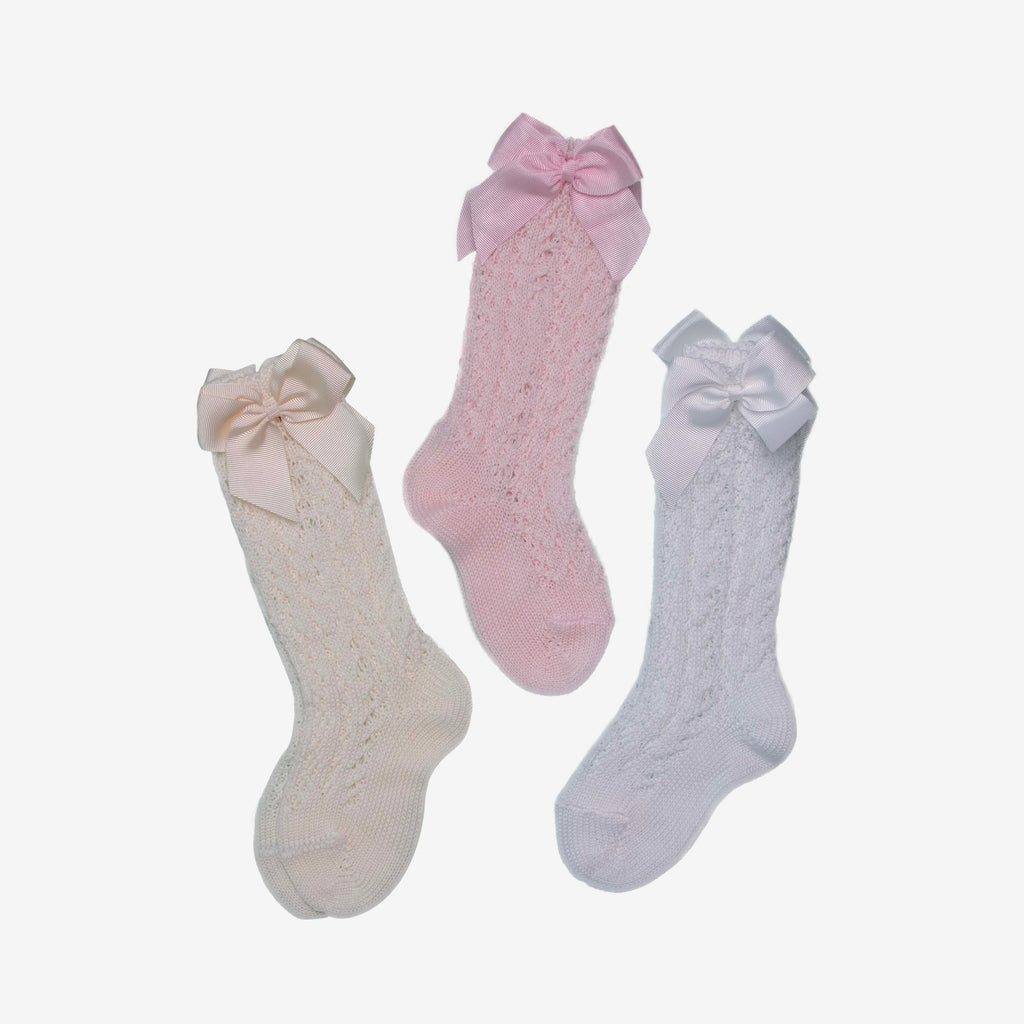 A studio product image displaying three pairs of knee-high baby socks in cream, light pink, and white. Each sock features an intricate pointelle lace knit pattern and is topped with a matching striped grosgrain ribbon bow (lazo) for a classic, vintage-inspired look.