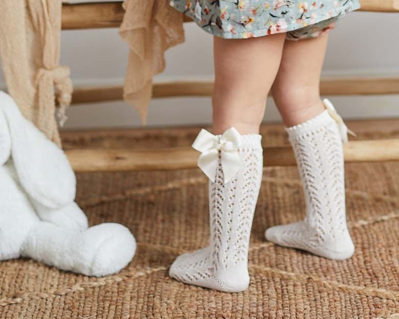 A lifestyle photograph of a baby’s legs from behind, wearing cream-colored knee-high socks with an intricate pointelle chevron lace knit. The socks feature a scalloped top edge and a matching ivory satin bow (lazo) on the outer side. The baby is standing on a natural jute rug next to a white plush bunny.