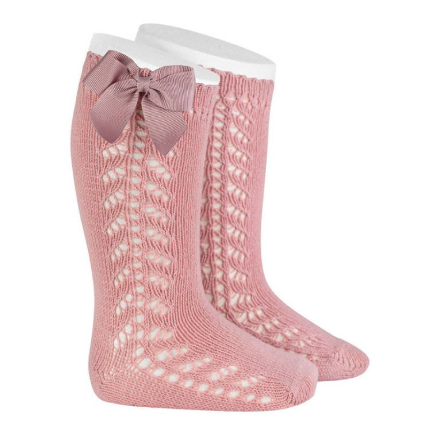 A studio product image of a pair of dusty rose pink knee-high baby socks. The socks feature a vertical chevron pointelle lace knit pattern, a scalloped top edge, and a matching mauve-pink grosgrain ribbon bow (lazo) on the side.