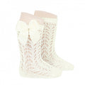 A studio product image of a pair of ivory white knee-high baby socks featuring an intricate, floral-inspired pointelle lace knit. The socks are finished with a scalloped top edge and a large, coordinating ivory satin bow (lazo) on the side.