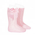 A studio product image of a pair of light pink knee-high baby socks featuring an intricate pointelle lace knit pattern. The socks are designed with a scalloped top edge and a large, coordinating pink grosgrain ribbon bow (lazo) stitched to the side.
