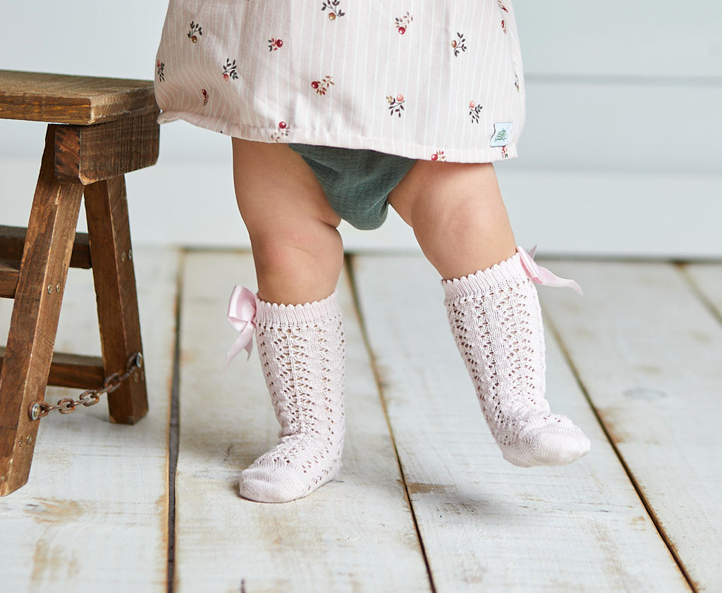 A close-up lifestyle shot of a baby’s legs wearing light pink knee-high socks with an intricate pointelle lace knit pattern. The socks feature a delicate scalloped edge at the top and are finished with a matching pink satin bow (lazo) on the side. The baby is standing on a white wooden floor next to a rustic wooden stool.