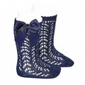 A lifestyle photograph of a baby’s legs wearing colored knee-high socks with a vertical pointelle lace knit pattern. The socks feature a delicate scalloped edge at the top and are finished with a matching navy blue satin bow (lazo) on the side. 