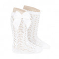A studio product image of a pair of bright white knee-high baby socks made from a delicate pointelle lace knit pattern. The socks feature a large, matching white grosgrain ribbon bow (lazo) on the side, displayed on light-colored forms against a plain white background.