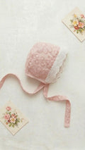 A flat lay of the Lola baby bonnet in a soft dusty pink, featuring intricate floral lace embroidery across the body. The bonnet is trimmed with a delicate white scalloped lace eyelet edge and has matching pink lace ties. It is styled on a neutral linen background with two vintage floral postcards.