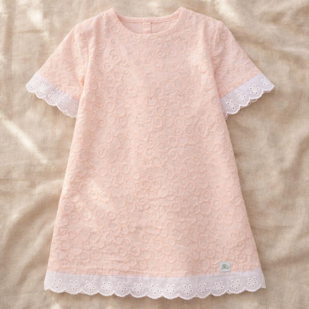 A detailed close-up of the Lola dress in dusty pink, showcasing the intricate floral lace embroidery and a delicate white eyelet lace trim along the hem. The dress is styled on a neutral linen backdrop with a vintage floral postcard, emphasizing its handmade, heirloom quality.