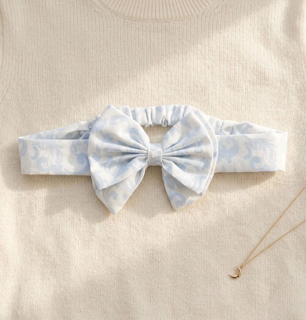 A flat lay photograph of the Malaga baby headband, featuring a soft white fabric with a light blue damask-style swirl pattern. The headband includes an elasticated back for comfort and a large matching fabric bow at the centre. It is styled on a cream-colored knitted sweater background next to a delicate gold moon pendant necklace.