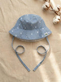 A top-down photograph of the Menorca baby bucket hat, featuring a light blue and white fine pinstriped fabric adorned with small embroidered white daisies. The hat includes matching pinstriped chin tie straps and a small square turtle brand label on the side. It is styled flat on a textured, neutral beige linen background alongside a natural cotton boll branch under soft sunlight.