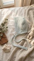 A side-view photograph of a light mint green baby bonnet made from a textured waffle-weave fabric. The bonnet features a wide, white scalloped eyelet lace trim and long matching mint waffle-knit ties. It is styled on a neutral linen backdrop next to a small knitted teddy bear, a pair of cream booties, and a leather notebook in soft, natural sunlight.