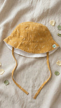 A flat lay photograph of a baby bucket hat made from mustard yellow muslin fabric with a white music note print. The hat features matching fabric chin straps and a small green turtle brand label on the brim. It is styled on a neutral linen backdrop with scattered white flower petals and green leaves.