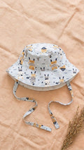 A flat lay photograph of a baby bucket hat featuring a light blue fabric with a pirate-themed print, including anchors, treasure maps, and stars. The hat has matching fabric chin straps and is styled on a neutral linen backdrop with a sprig of dried wheat in the lower right corner.