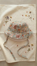 A flat lay photograph of the Primavera baby bucket hat in a soft sage green fabric with a white paisley and floral pattern. The hat features a matching chin strap and a small green turtle brand label on the brim. It is styled on a neutral linen surface next to a sprig of dried lavender and a small stone in soft, natural light.