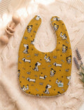 A flat lay of a mustard yellow baby bib featuring a whimsical pattern of various cartoon puppies, tiny bones, and paw prints. The bib has white snap closures at the top and is resting on a soft pink cable-knit sweater alongside a wooden rattle and lavender sprigs.