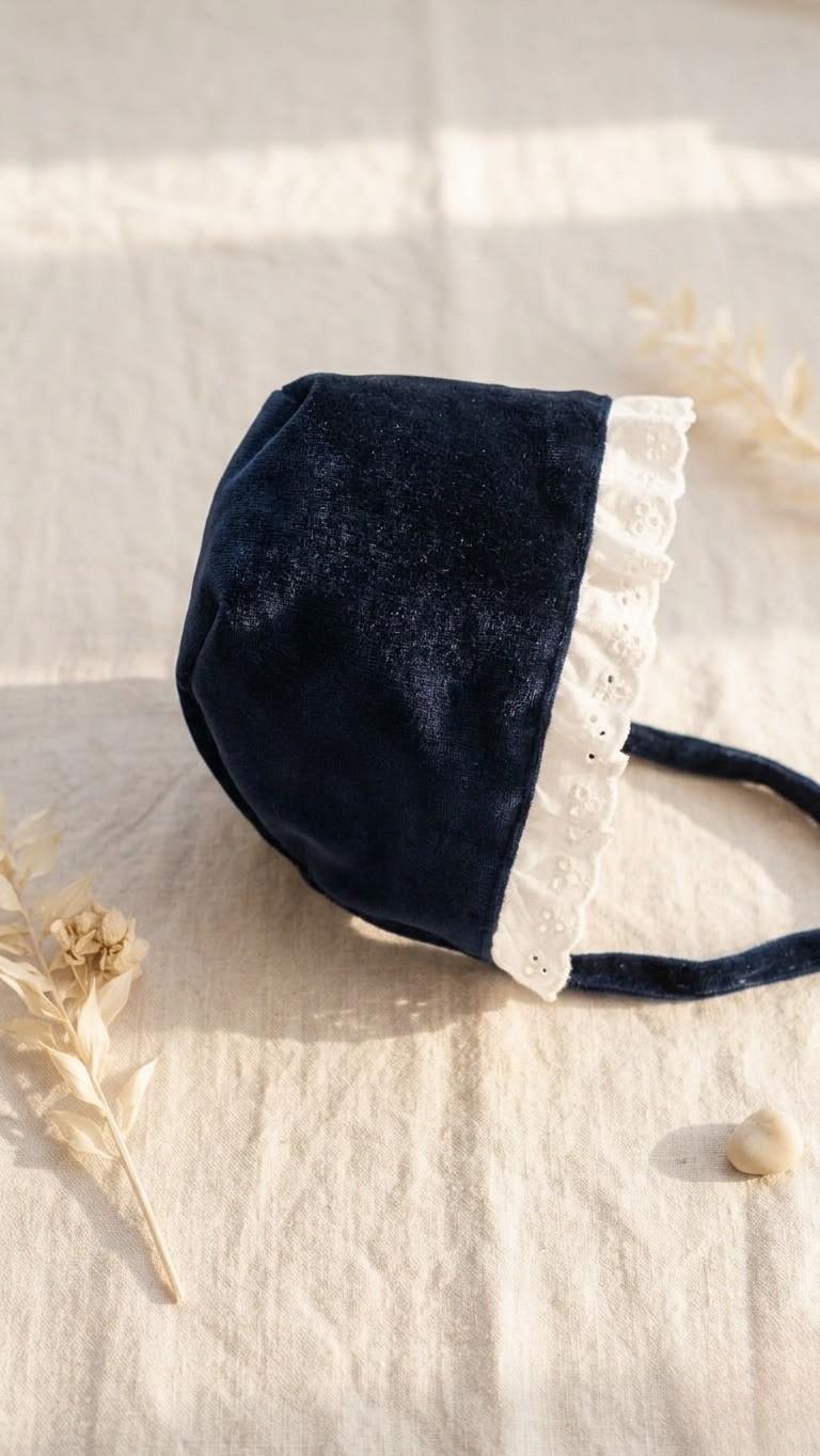 A side-profile photograph of a royal blue velvet baby bonnet, showcasing its plush texture and rounded shape. The bonnet is finished with a crisp white eyelet lace trim and matching velvet ties, resting on a neutral linen surface with soft shadows and dried floral accents.