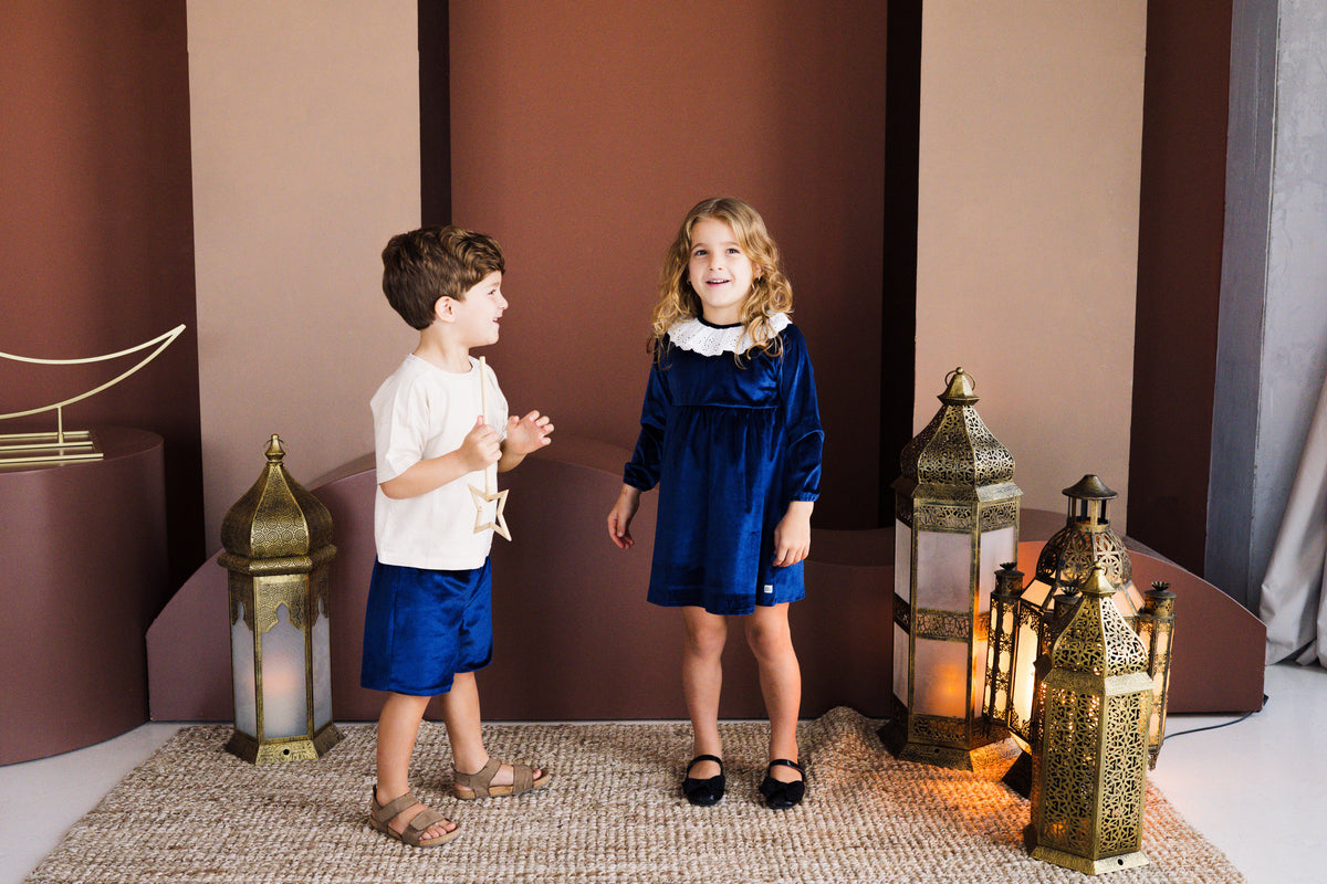 A studio lifestyle photograph of a young boy and girl posing together on a woven rug. The girl stands smiling in a long-sleeved royal blue velvet dress with a white ruffled lace collar and black Mary Jane shoes. The boy stands beside her holding a star wand, wearing royal blue velvet shorts, a white t-shirt, and brown sandals. They are surrounded by ornate brass lanterns against a warm-toned backdrop.