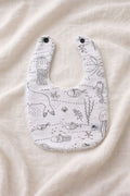A flat lay photograph of a handmade baby bib featuring a white fabric with black line-art illustrations of undersea life, including jellyfish, seahorses, fish, and coral. The bib has two black snap fasteners at the neck and a soft white towelling texture visible along the edges. It is styled against a neutral, draped cream-colored fabric background.