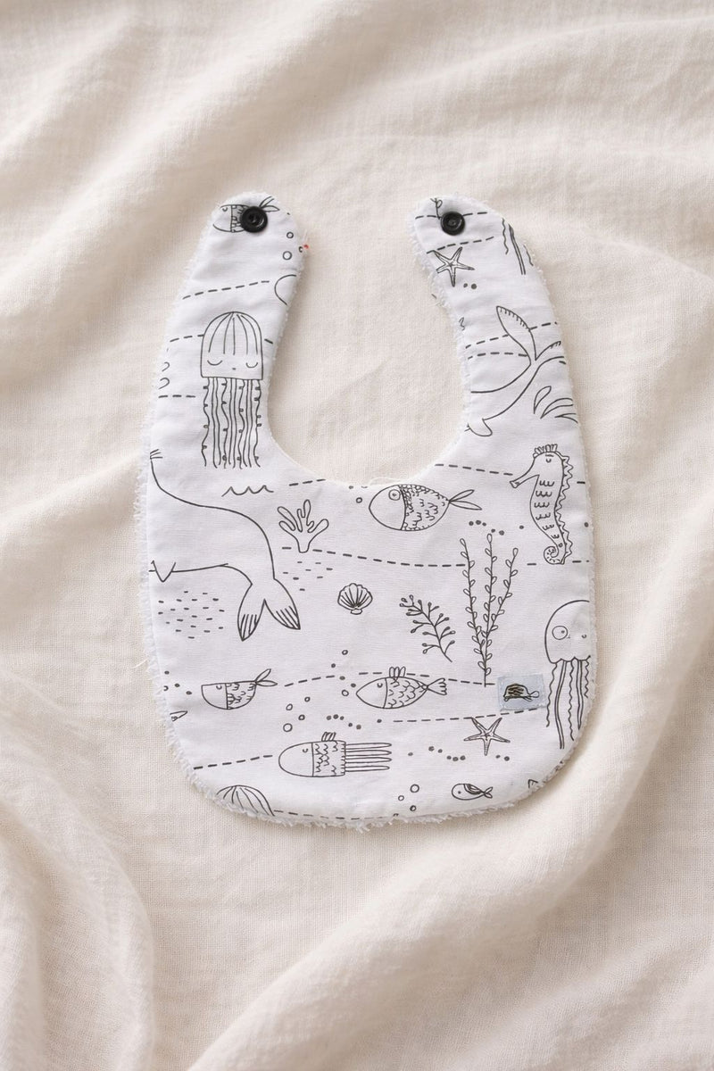 A flat lay photograph of a handmade baby bib featuring a white fabric with black line-art illustrations of undersea life, including jellyfish, seahorses, fish, and coral. The bib has two black snap fasteners at the neck and a soft white towelling texture visible along the edges. It is styled against a neutral, draped cream-colored fabric background.