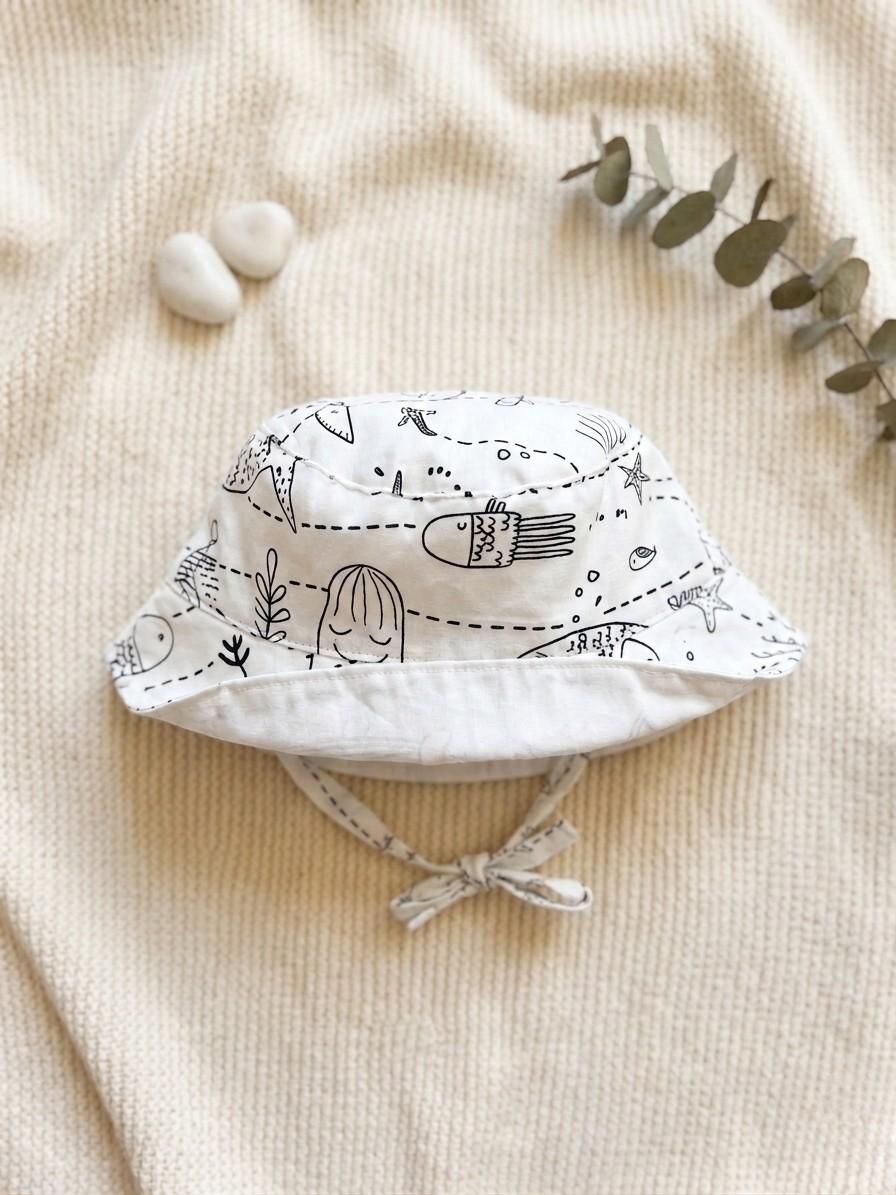 A flat lay photograph of a handmade baby bucket hat featuring a white fabric with black line-art illustrations of sea creatures, including fish, starfish, and seaweed. The hat has a matching fabric chin strap tied in a bow and is styled on a cream knitted blanket next to two small white stones and a sprig of eucalyptus.