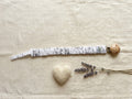 A flat lay photograph of the Under the Sea pacifier clip, featuring a white fabric strap with black line-art illustrations of ocean life. The strap has a ruffled, elasticated texture and is attached to a round, natural light-wood clip. It is styled on a neutral, textured cream fabric background alongside a small white felt heart and sprigs of dried lavender.