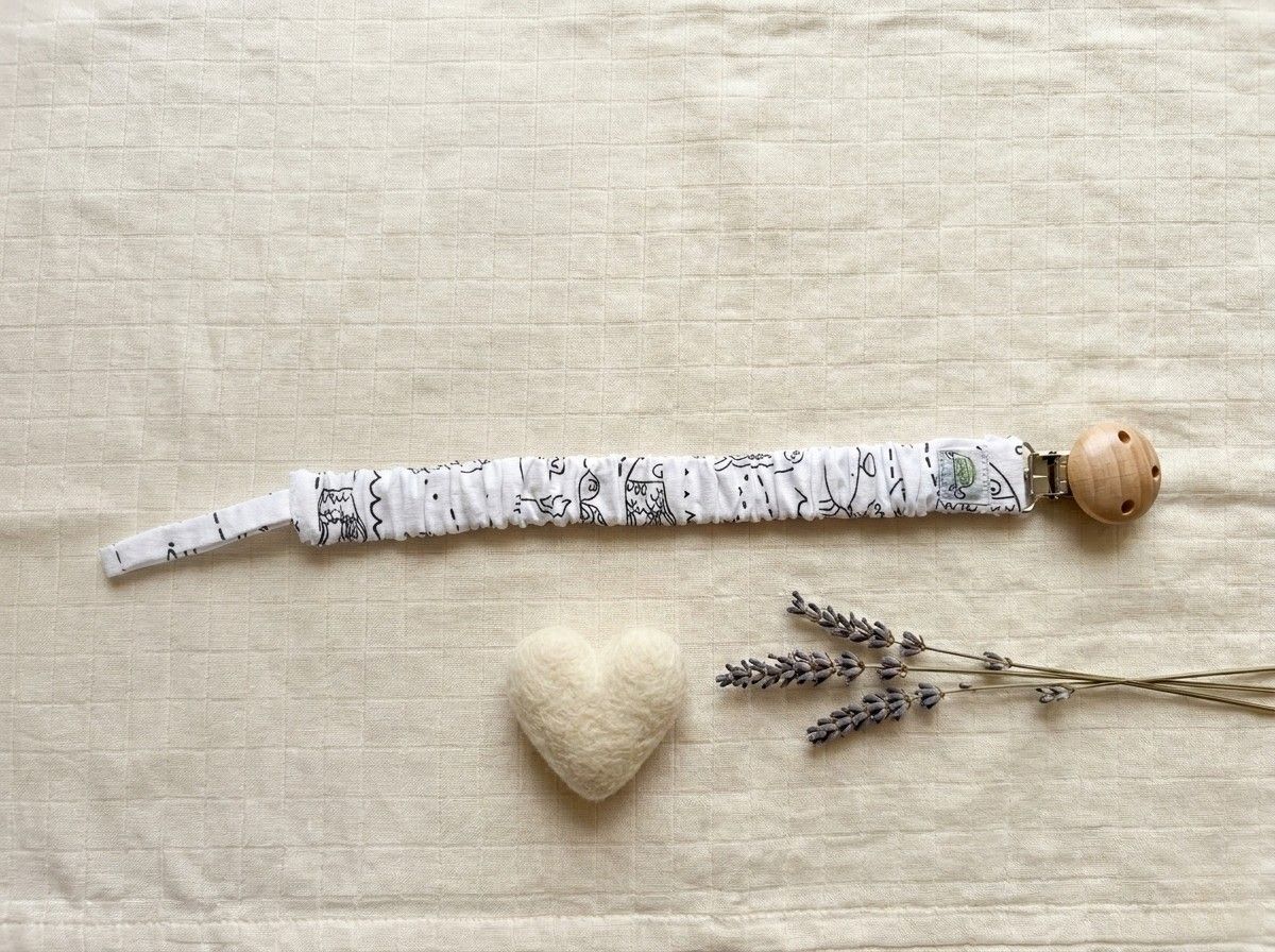 A flat lay photograph of the Under the Sea pacifier clip, featuring a white fabric strap with black line-art illustrations of ocean life. The strap has a ruffled, elasticated texture and is attached to a round, natural light-wood clip. It is styled on a neutral, textured cream fabric background alongside a small white felt heart and sprigs of dried lavender.