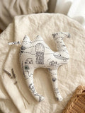 A handmade camel-shaped plush cushion made from white fabric with a black line-art "Under the Sea" print featuring a jellyfish, whale, sea lion, and coral. The cushion is styled on a neutral linen background with a sprig of dried lavender and the corner of a wicker basket, creating a soft, minimalist nursery aesthetic.