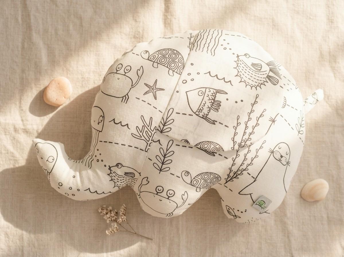 A handmade elephant-shaped plush cushion made from white fabric with a black line-art "Under the Water" print. The design features various sea creatures, including a whale, jellyfish, and sea lion, meant for colouring. The cushion is styled on a neutral linen backdrop with a sprig of dried white flowers and couple seashells on the sides.