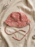 A flat lay of the Verano baby bucket hat in a warm terracotta orange, featuring a white paisley and floral pattern organic cotton muslin fabric. The hat includes matching chin ties and a small green turtle brand logo on the brim. It is styled on a neutral linen fabric with a sprig of dried lavender and a small smooth stone.