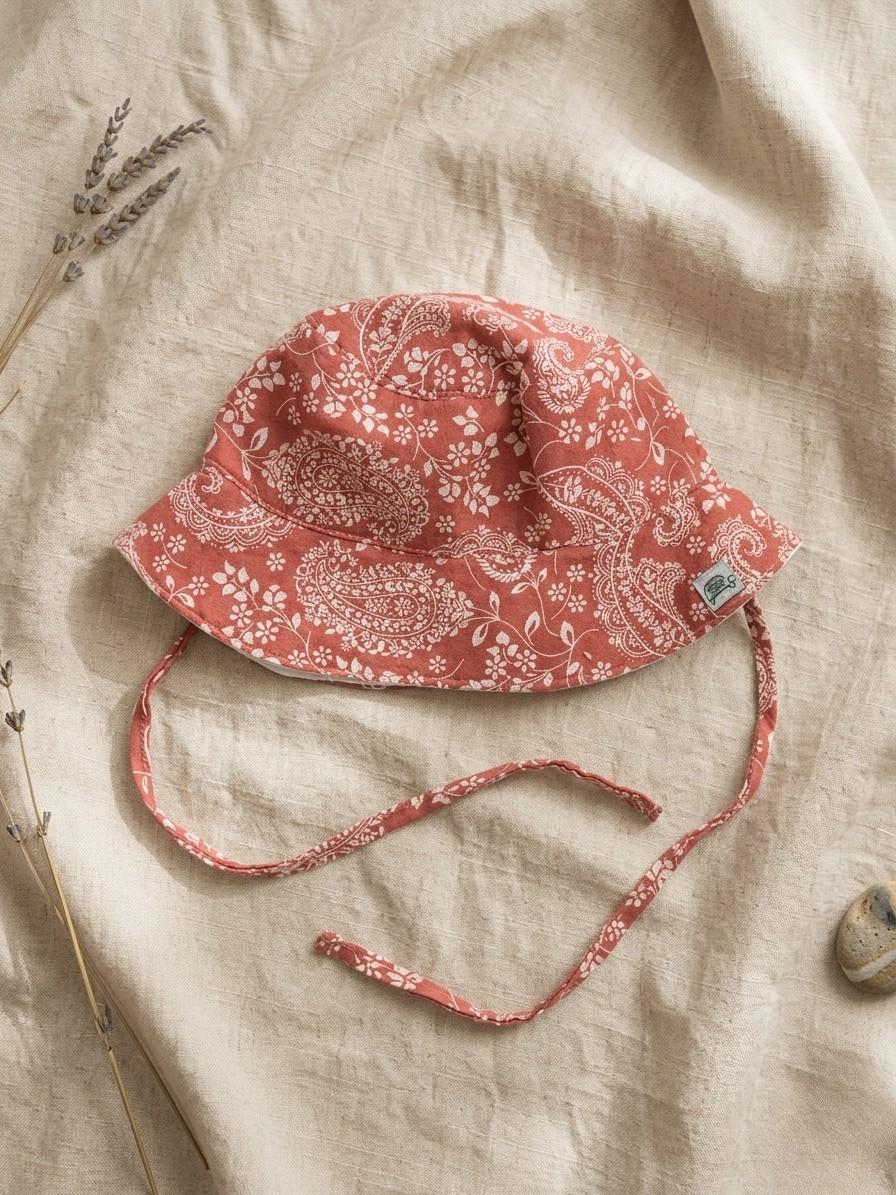 A flat lay of the Verano baby bucket hat in a warm terracotta orange, featuring a white paisley and floral pattern organic cotton muslin fabric. The hat includes matching chin ties and a small green turtle brand logo on the brim. It is styled on a neutral linen fabric with a sprig of dried lavender and a small smooth stone.