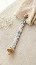A flat lay photograph of a handmade fabric pacifier clip with a vintage car and vehicle print on a white background. The clip features a natural wooden fastener and holds a tan-colored pacifier. It is styled on a neutral linen surface next to a cream knitted blanket and a delicate sprig of white baby's breath flowers.