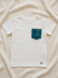 A flat lay photograph of a crisp white, short-sleeved cotton t-shirt for children. The shirt features a single chest pocket made from textured emerald green velvet fabric and a small grey brand label at the bottom hem. It is styled against a neutral, cream-colored draped fabric background.
