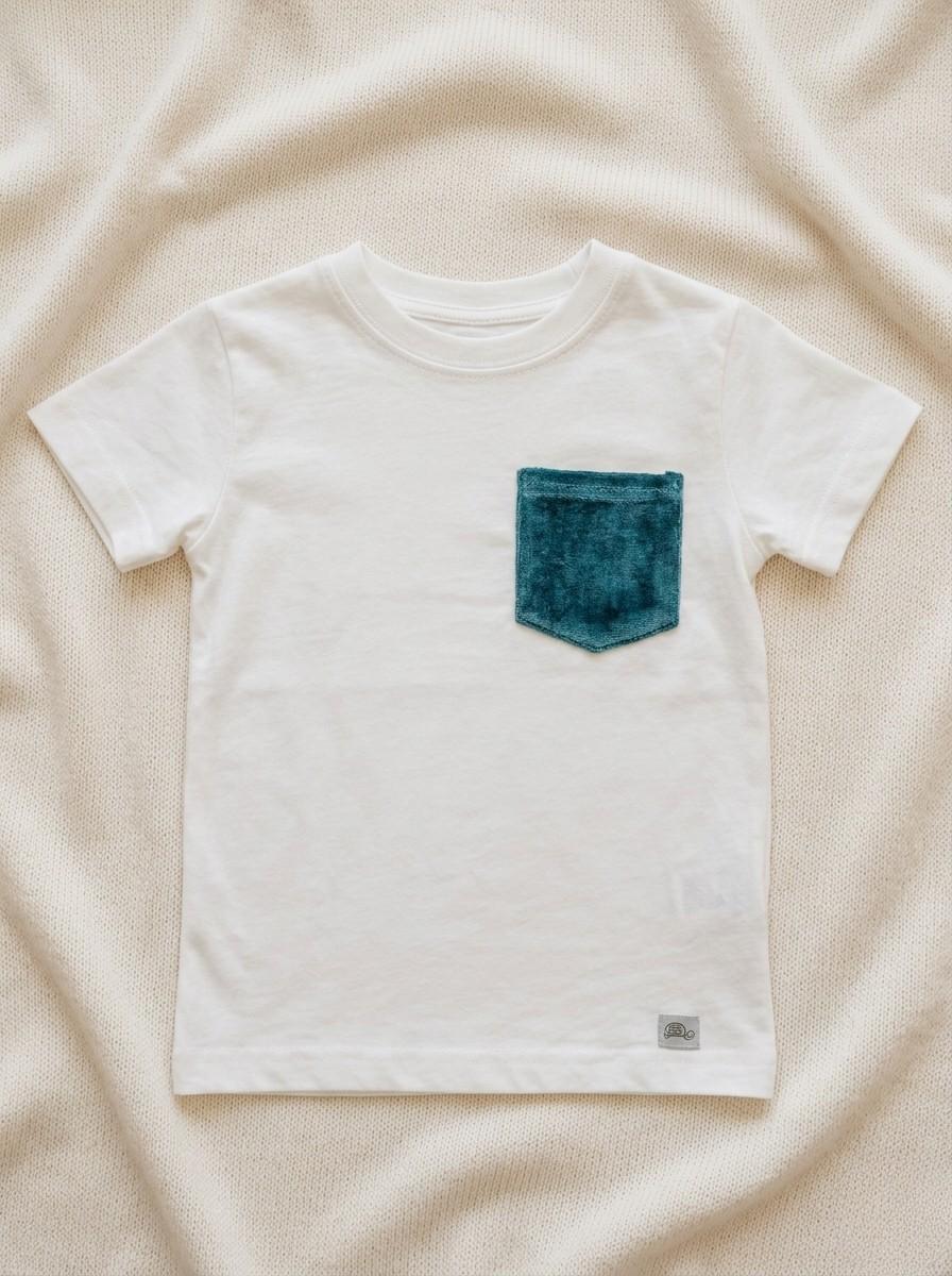 A flat lay photograph of a crisp white, short-sleeved cotton t-shirt for children. The shirt features a single chest pocket made from textured emerald green velvet fabric and a small grey brand label at the bottom hem. It is styled against a neutral, cream-colored draped fabric background.