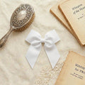 A flat lay photograph of a crisp white grosgrain ribbon hair bow with long tails. The bow is centered on a neutral linen fabric, surrounded by vintage aesthetic props including an ornate silver hairbrush and antique books.