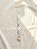A flat lay photograph of the Zoo pacifier clip, featuring a fabric strap with a white base and a colourful illustration of various zoo animals. The strap has a ruffled, elasticated texture and is attached to a round, natural light-wood clip. It is styled vertically on a neutral, draped cream linen background in soft sunlight.