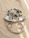 A top-down photograph of a baby bucket hat featuring a light grey or beige fabric covered in a playful zoo animal print, including a zebra, tiger, alligator, and elephant. The hat includes matching fabric chin tie straps and is styled flat on a neutral, textured beige linen background under soft, warm sunlight.
